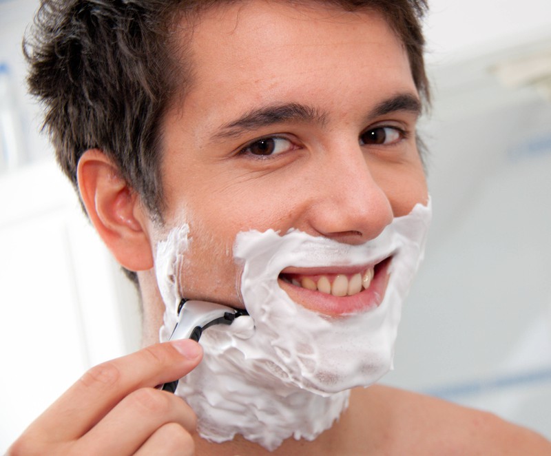 Junger Mann rasiert sich mit Rasierklinge und Rasierschaum im Badezimmer um gepflegt zu wirken